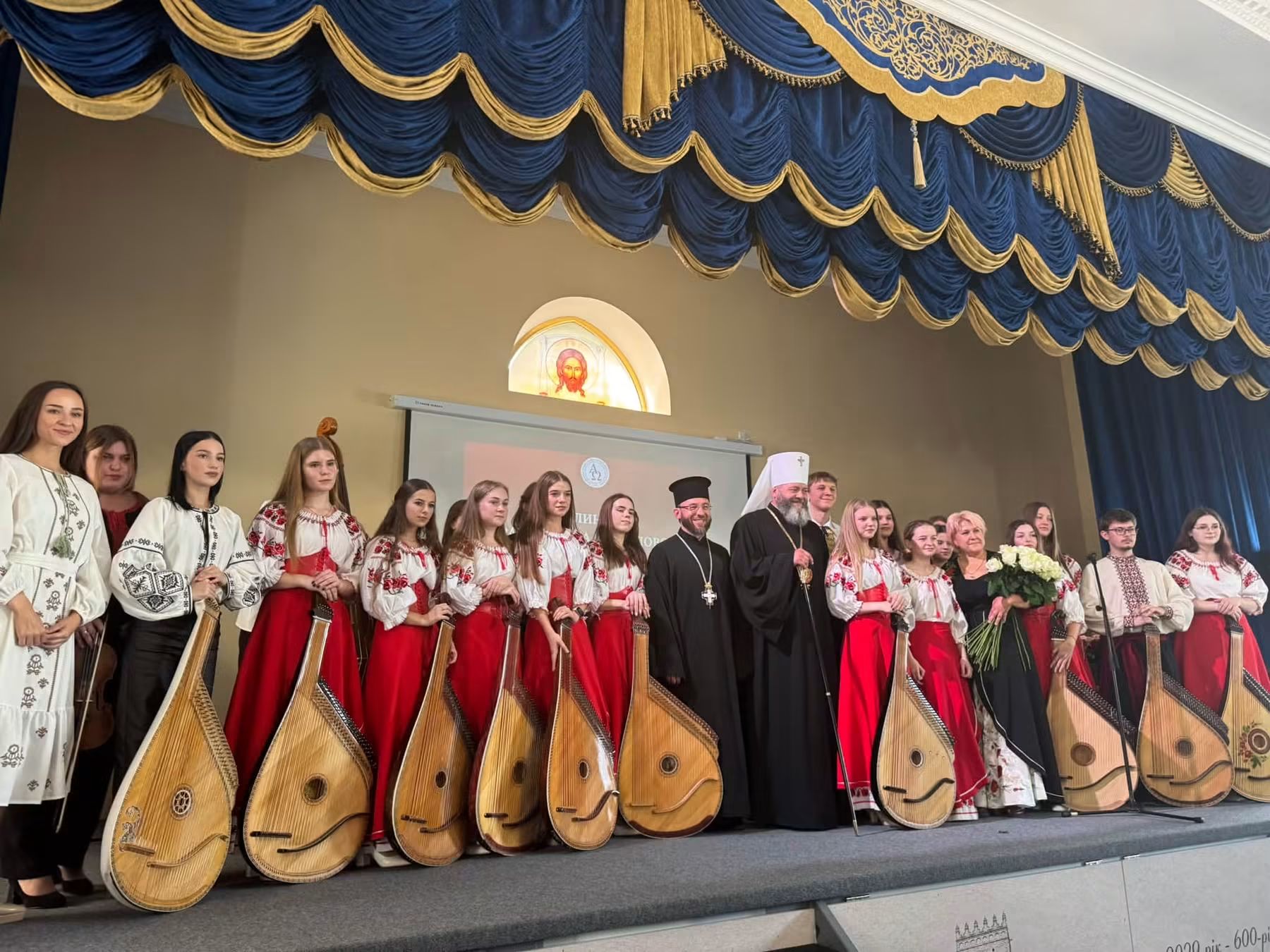 Дівчата у вишиванках і червоних спідницях із бандурами стоять на сцені разом з архієреєм у білому клобуку, священником і жінкою з букетом білих троянд; над сценою вітраж із Нерукотворним образом Спасителя
