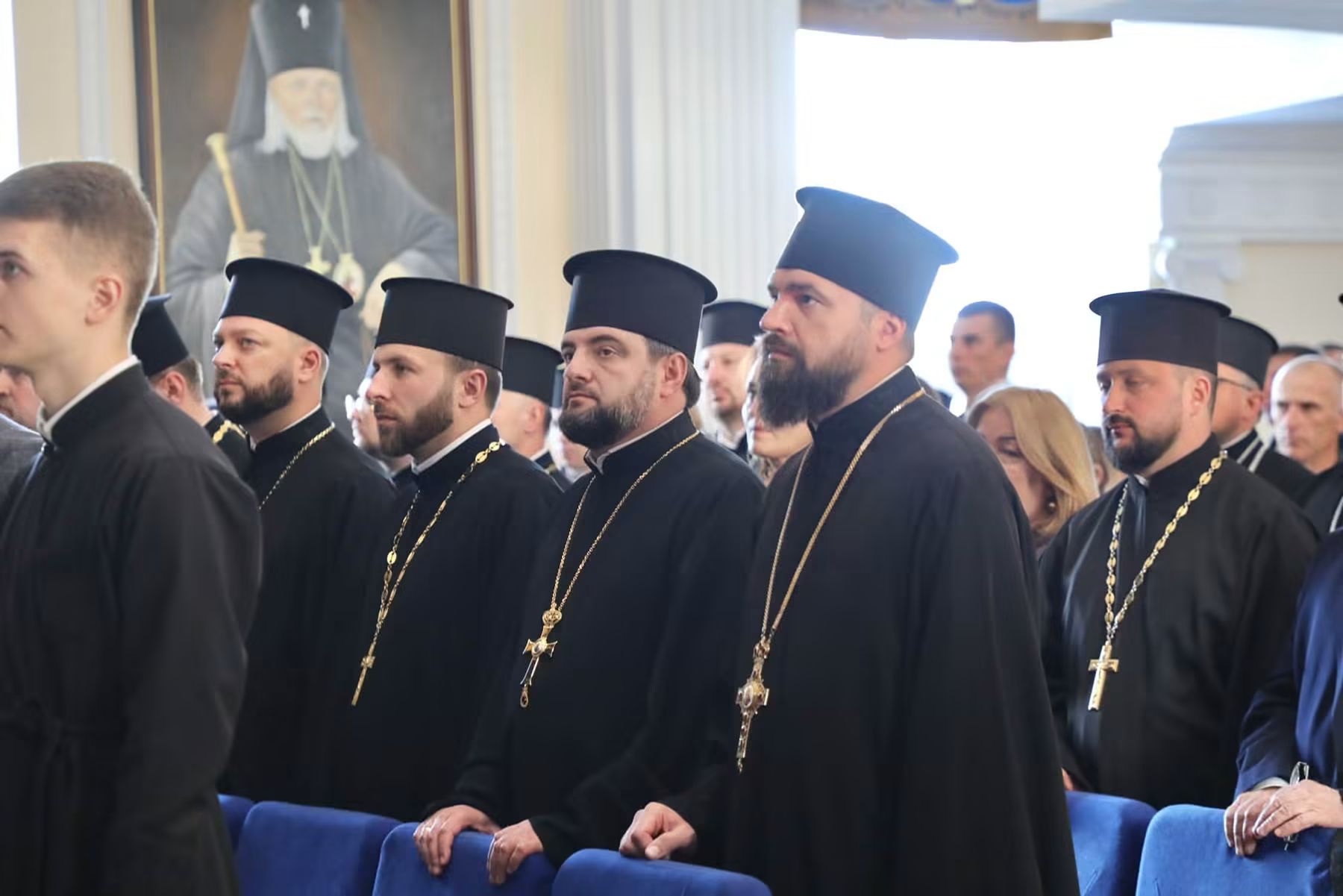 Священники в чорних підрясниках і камилавках з наперсними хрестами стоять у залі між синіх крісел; позаду портрет архієрея в білому клобуку