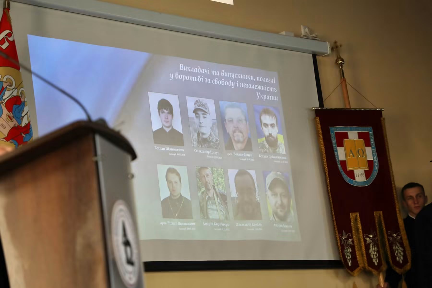 На екрані в актовій залі слайд «Викладачі та випускники, полеглі у боротьбі за свободу і незалежність України» з вісьмома портретами; праворуч хоругва академії