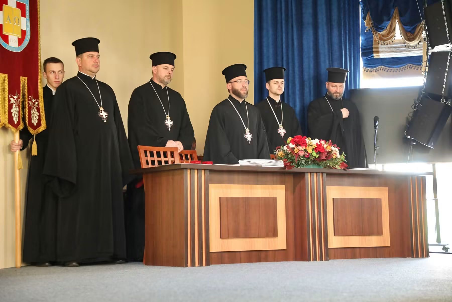 П’ятеро священників у чорних підрясниках і камилавках із наперсними хрестами стоять за столом президії з квітами; ліворуч студент тримає хоругву академії з літерами Альфа і Омега та датою 1796