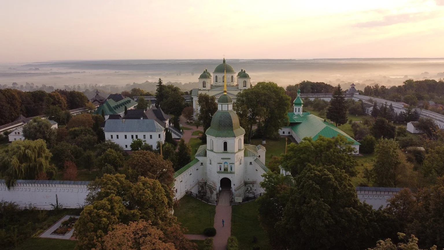 Вигляд згори на білий монастир із зеленими банями: надбрамна дзвіниця з позолоченим шпилем, за нею п'ятибанний собор, довкола мури й дерева, удалині туман над рікою