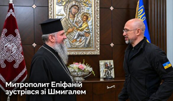 Митрополит Епіфаній у чорному клобуку розмовляє з Денисом Шмигалем у темній куртці з шевроном прапора України; позаду ікона Богородиці з Дитям у срібному окладі, хоругва і прапор України; напис «Митрополит Епіфаній зустрівся зі Шмигалем»