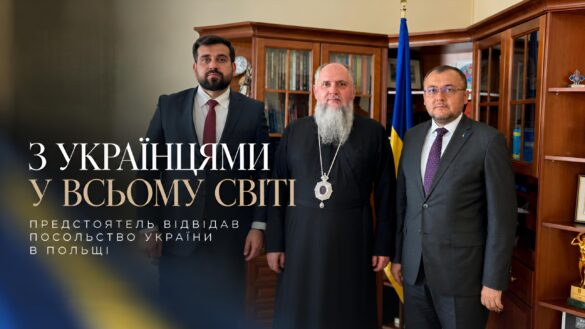 Православний архієрей у чорному підряснику з панагією і двоє чоловіків у темних костюмах стоять у кабінеті з книжковими шафами біля державного прапора України; напис «З українцями у всьому світі. Предстоятель відвідав Посольство України в Польщі»